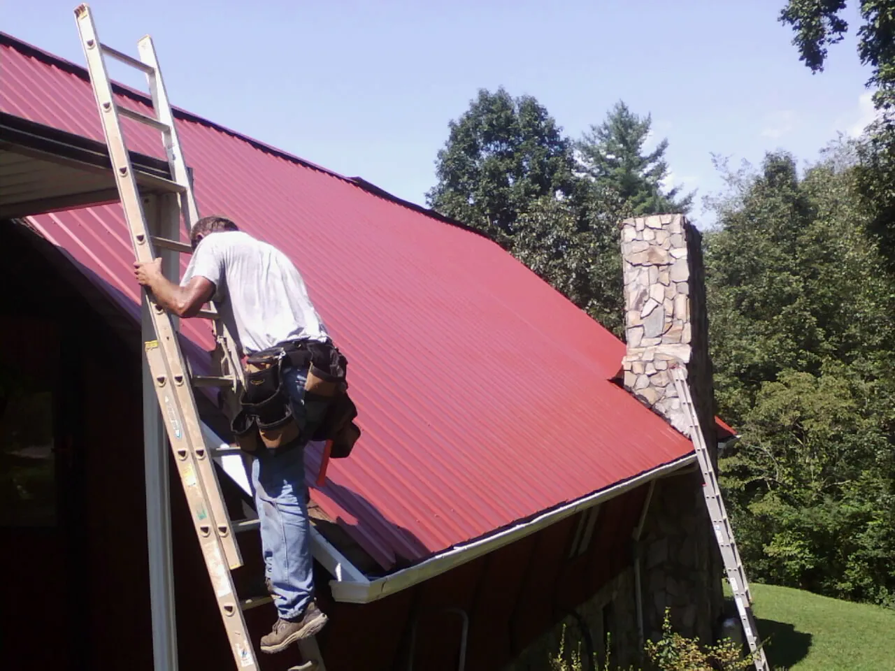 Roofer on a ladder beside a red metal roof and stone chimney — Soffit & Fascia Repair by Roofing Brothers in Goldvein